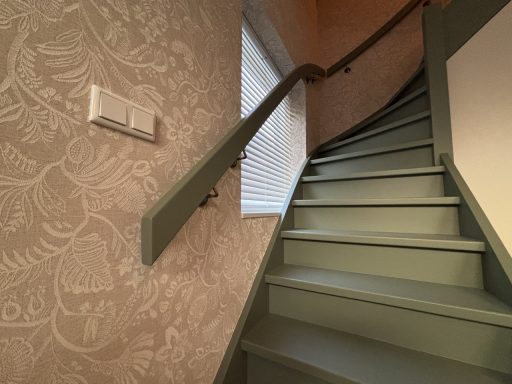 Groene trap met leuning, tegen een muur met behang en een raam aan de bovenkant.